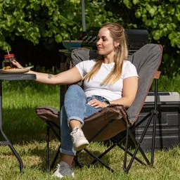 Person sitzt entspannt in einem Campingstuhl, hält einen Teller und ein Glas mit Strohhalm. Grünes Gras im Hintergrund.