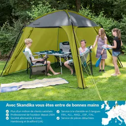 Zelt im Freien mit mehreren Personen, die in Stühlen sitzen. Auf einem Tisch stehen Geschirr und Getränke. Grüne Wiese im Hintergrund.