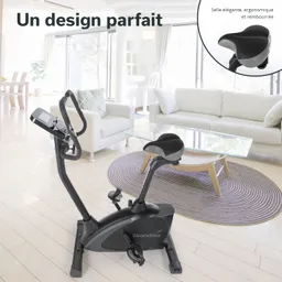 Ein schwarzes Ergometer steht in einem hellen Raum mit Holzfußboden. Im Hintergrund sind ein Sofa, ein Tisch und ein Fenster sichtbar.