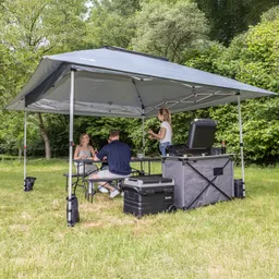 Zelt mit grauem Dach, darunter drei Personen an einem Tisch, eine weitere Person steht. Kühlbox und Grill sichtbar. Grünes Umfeld.