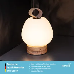 Tischlampe mit milchig-weißer Glaskuppel und Holzfuß. Oben mit einem Lederriemen zum Tragen. Heller, neutraler Hintergrund.