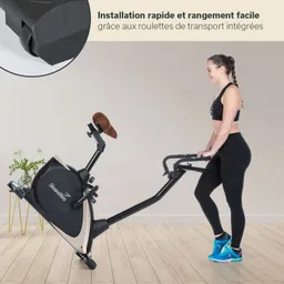 Eine Sportlerin in enger Kleidung steht neben einem Fitnessgerät. Das Gerät hat eine schwarze und goldene Farbe und ist auf einem Holzboden platziert. Im Hintergrund sind eine Pflanze und ein kleiner Tisch zu sehen.