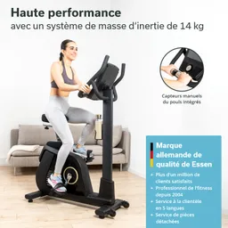 Eine Person sitzt auf einem Heimtrainer in einem hellen Raum. Der Trainer hat ein schwarzes Design, und die Person trägt sportliche Kleidung. Im Hintergrund ist ein Sofa sichtbar.