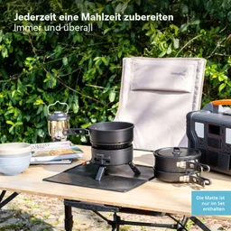 Kochutensilien auf einem Tisch im Freien: ein Kocher, zwei Töpfe, Schalen und eine Laterne, umgeben von grünen Pflanzen.