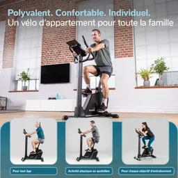 Stationäres Fitnessgerät in einem Raum mit großen Fenstern. Eine Person sitzt aktiv auf dem Fahrrad. Im Hintergrund sind Pflanzen und ein Fernseher sichtbar.