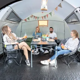 Vier Personen sitzen in einem Zelt um einen Tisch mit Snacks und Getränken. Bunte Wimpel sind dekorativ aufgehängt.