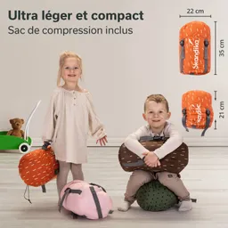 Zwei Kinder stehen in einem hellen Raum mit Rucksackmodellen. Eines der Kinder hält einen orangefarbenen Rucksack, während das andere auf einem grünen Kissen sitzt und einen braunen Rucksack umarmt. Weitere Rucksäcke liegen auf dem Boden.