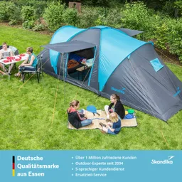 Zelt in der Natur mit mehreren Personen, die an einem Tisch sitzen und am Boden spielen. Grünes Gras und Bäume im Hintergrund.