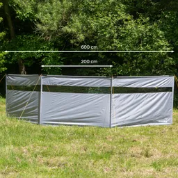 Graue Trennwand mit zwei sichtbaren Fenstern, auf einer Wiese, umgeben von Bäumen. Maße: 600 cm Breite, 200 cm Höhe.