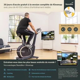 Person sitzt auf einem Heimtrainer vor einem großen Bildschirm, der eine Radfahrstrecke zeigt. Im Hintergrund Fenster mit Blick ins Freie.