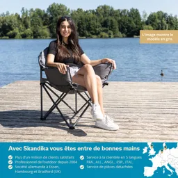 Person sitzt auf einem grauen Klappstuhl an einem Gewässer, umgeben von grünem Ufer und Bäumen.