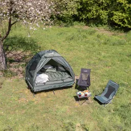 Campingzelt auf einer Wiese mit zwei Stühlen und einem Tisch, auf dem Snacks und Getränke stehen. Blühender Baum im Hintergrund.