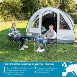 Zelt im Freien mit zwei Personen, die an einem Tisch mit Obst und Getränken sitzen. Grüner Rasen und Bäume im Hintergrund.