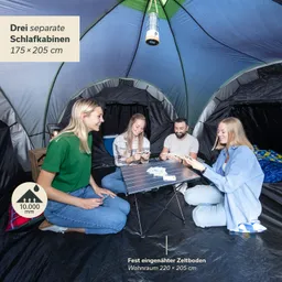 Vier Personen sitzen um einen kleinen Tisch in einem großen Zelt. Der Zeltinnenraum ist farbenfroh und bietet drei Schlafkabinen. Ein hängendes Licht ist sichtbar.