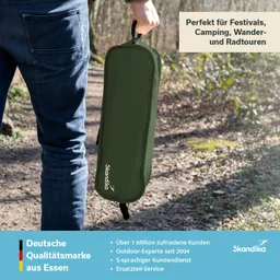 Eine Person hält eine grüne Tasche mit dem Logo "Skandika" in einer natürlichen Umgebung. Der Weg ist schmal und von Bäumen umgeben.