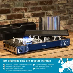 Gasherd in blauer Farbe mit brennender Flamme, Holzunterlage, Metallplatte im Hintergrund und schwarzem Koffer. Steinwand im Hintergrund.