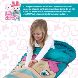 Ein Kind in einem weißen Shirt sitzt in einem Schlafsack mit einem bunten, cartoonhaften Gesicht. Der Hintergrund zeigt ein Muster aus Blumen und Punkten.
