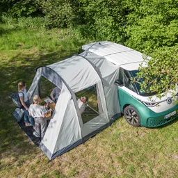 Zelt neben einem VW-Fahrzeug auf einer Wiese. Drei Personen stehen am Tisch, umgeben von Bäumen.