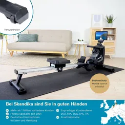 Rudergerät auf einer Schutzmatte in einem hellen Raum. Im Hintergrund eine couch und ein Beistelltisch. 횡부분 zeigt Transportrollen.