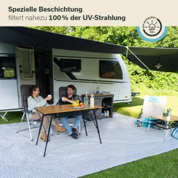Ein Wohnwagen steht auf einem grünen Rasen. Vor dem Wohnwagen sitzen zwei Personen an einem Tisch, umgeben von Stühlen. Auf dem Tisch sind Obst und Getränke platziert. Eine große Markise bietet Schatten. Im Hintergrund sind Bäume sichtbar.