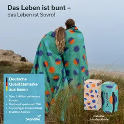 Zwei Personen stehen auf einem Strand, umhüllt von bunten Decken mit großen Punkten. Im Hintergrund sind Dünen und das Meer sichtbar. Neben ihnen steht eine Verpackung mit Schlafsack.