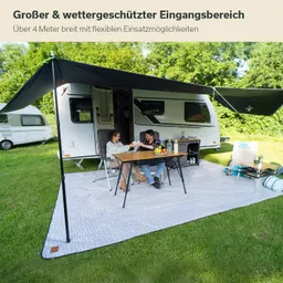 Ein Wohnmobil mit ausgeklappter Markise steht auf einer grünen Wiese. Zwei Personen sitzen an einem Tisch, auf dem Obst und Getränke stehen. Das Bodenlager ist mit einem grauen Teppich bedeckt.
