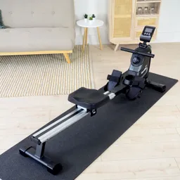 Rudergerät auf einer schwarzen Unterlage, mit Sitz, Fußstützen und einem Display. Im Hintergrund Sofa und kleiner Tisch.