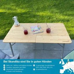 Tisch aus Holz mit zwei Gläsern, einem Magazin und einer Laterne auf grüner Wiese. Stuhl im Hintergrund.
