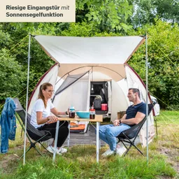 Zwei Personen sitzen an einem Tisch vor einem Zelt. Im Hintergrund sind Bäume zu sehen, und das Zelt hat ein großes Eingangstor mit Sonnensegel.