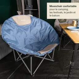 Ein blauer, rundlicher Campingsessel mit einem grauen Polster an der Rückenlehne steht auf einem schwarzen Untergrund. Im Hintergrund sind weitere Möbel und ein Tisch mit Geschirr sichtbar.