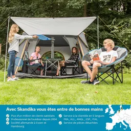 Familie in einem Zelt auf einer Wiese, zwei Personen sitzen, eine Person liest, eine Person steht mit einem Schläger.