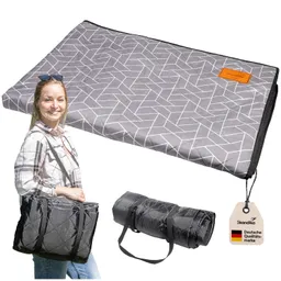 Faltbare Picknickdecke in Grau mit geometrischem Muster, daneben eine Person in kariertem Hemd mit Tasche und gerollte Decke.