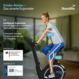 Ergometriegerät in einem hellen Raum, eine Person sitzt darauf, lächelt und trägt Sportkleidung. Pflanzen im Hintergrund.