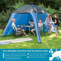 Zelt in Blau und Grau im Freien, umgeben von Bäumen. Vier Personen sitzen und stehen im Zelt, mit einem Tisch und Stühlen.