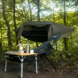 Ein Campingplatz im Wald mit einem hängenden Zelt, einem klappbaren Tisch, einer Lampe, einer Wasserflasche und einem Stuhl.