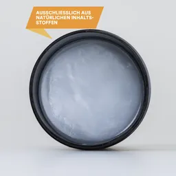 Runder Behälter mit grauer Creme oder Paste, auf einem hellen Hintergrund, mit Hinweistext in Orange oben links.