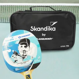 Tischtennisschläger mit einer blauen Oberfläche und der Aufschrift „Joey Kelly LEVEL 500“ neben einer schwarzen Tasche mit dem Logo „Skandika by Schildkröt“. Im Hintergrund ist ein Tischtennisnetz zu sehen.
