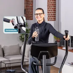 Eine Person steht an einem elliptischen Trainer, hält eine Wasserflasche in der Hand und lächelt. Im Hintergrund sind ein Sofa und eine Pflanze zu sehen.