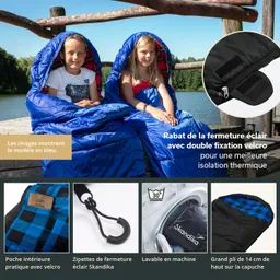 Zwei Kinder sitzen auf einer Holzplanke am Wasser, in blauen Schlafsäcken. Sie lächeln in die Kamera.