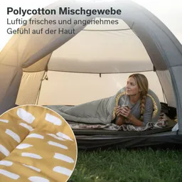 Zeltinneneinstellung mit Person, die in einem Schlafsack liegt und eine Tasse hält. Im Vordergrund ein Stoffmuster mit gelben und weißen Elementen.