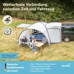 Zelt mit Anbau, Verbindung zu einem Fahrzeug. Zwei Personen sitzen an einem Tisch. Grüne Wiese, Bäume im Hintergrund.
