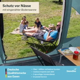Ein Mann sitzt entspannt in einem Faltstuhl, während zwei Kinder auf einer Decke spielen. Ein Tisch mit Getränken und einem Buch ist im Vordergrund sichtbar.