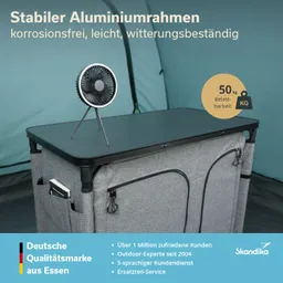 Ein stabiler Aluminiumrahmen mit einem schwarzen Tischoberfläche. Ein kleiner Ventilator steht darauf. Der Unterbau ist grau mit zwei Taschen.