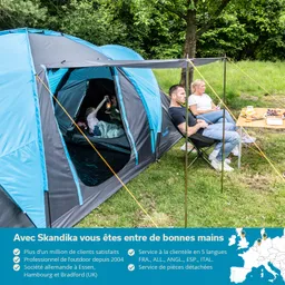 Zelt mit blauen und grauen Elementen, eine Person mit einer Lampe, zwei sitzende Personen neben einem Tisch mit Essen.