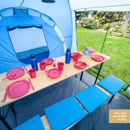 Campingplatz mit blauer Zeltkonstruktion und einem Tisch aus Holz. Darauf rote Teller, Tassen und Besteck sowie blaue Sitzkissen.
