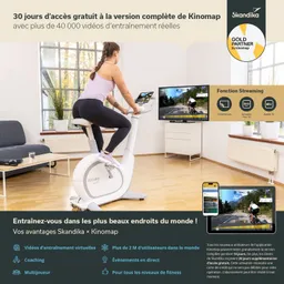 Eine Person trainiert auf einem Heimtrainer in einem hellen Raum. Im Hintergrund ist ein Fernseher mit einer Trainingsvideo-Anzeige. Ein Tablet mit der gleichen Anzeige steht auf dem Tisch.