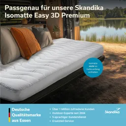 Ein gepolstertes Bett im Inneren eines Fahrzeugs, mit weißen und orangenen Kissen. Im Hintergrund Natur mit Wasser.