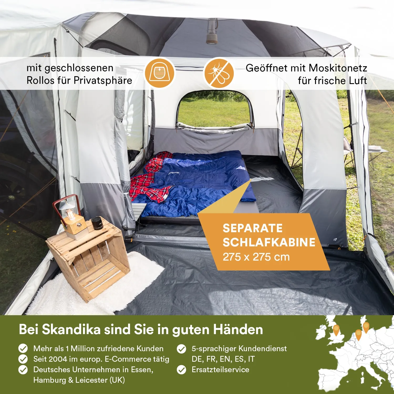 Zeltinterieur mit Schlafbereich, Schlafsäcken und Holztisch. Fenster mit Moskitonetz und geschlossenen Rollos. Grüne Wiese außen.