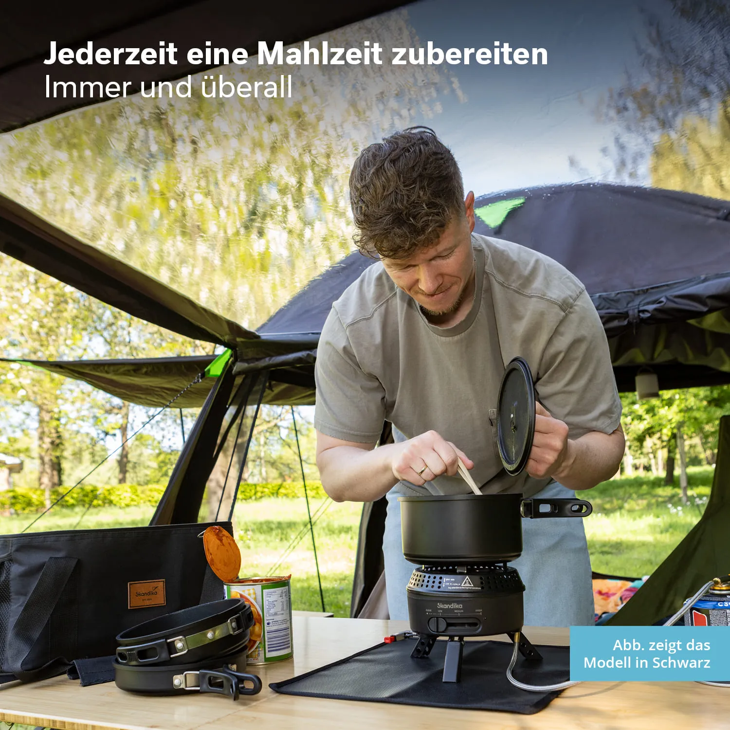 Person kocht am Campingtisch mit einem tragbaren Kocher. Darunter befindet sich ein schwarzer Topf. Im Hintergrund ist ein Zelt.