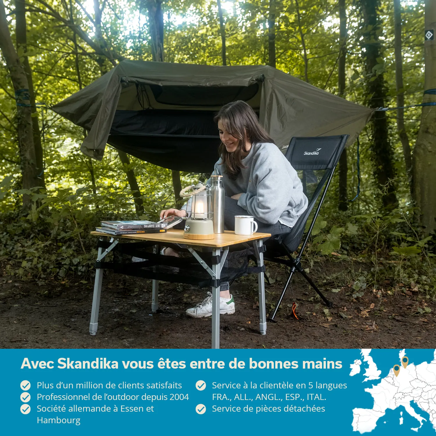 Person sitzt an einem Campingtisch im Wald, neben einer Hängematte. Auf dem Tisch stehen eine Lampe, Bücher und eine Tasse.